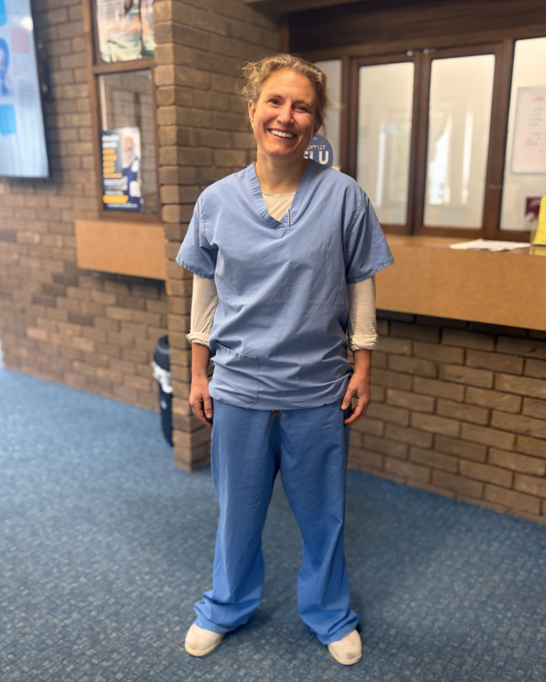 A doctor wearing light blue scrubs stands in a corridor with clue carpet and brick walls
