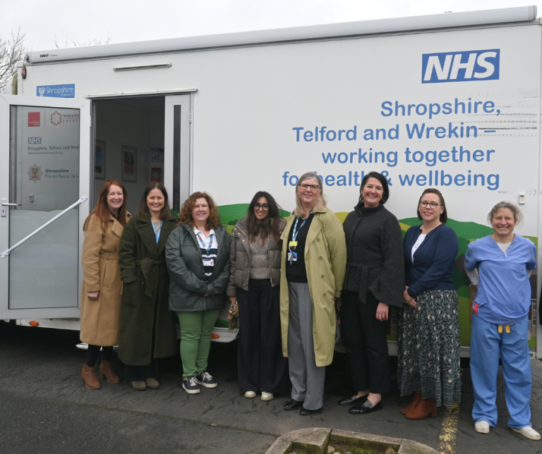 group of people wearing coats stand alongside a large white van. The van has an illustration of rural landscape on the side and a blue NHS logo