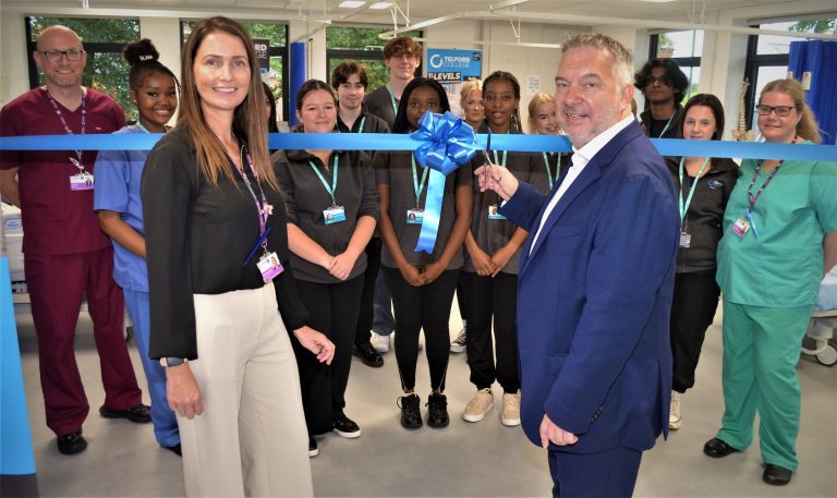 A man in a suit cuts a blue ribbon. There is a woman standing next to him and behind the ribbon there are people in NHS uniform looking towards the camera.